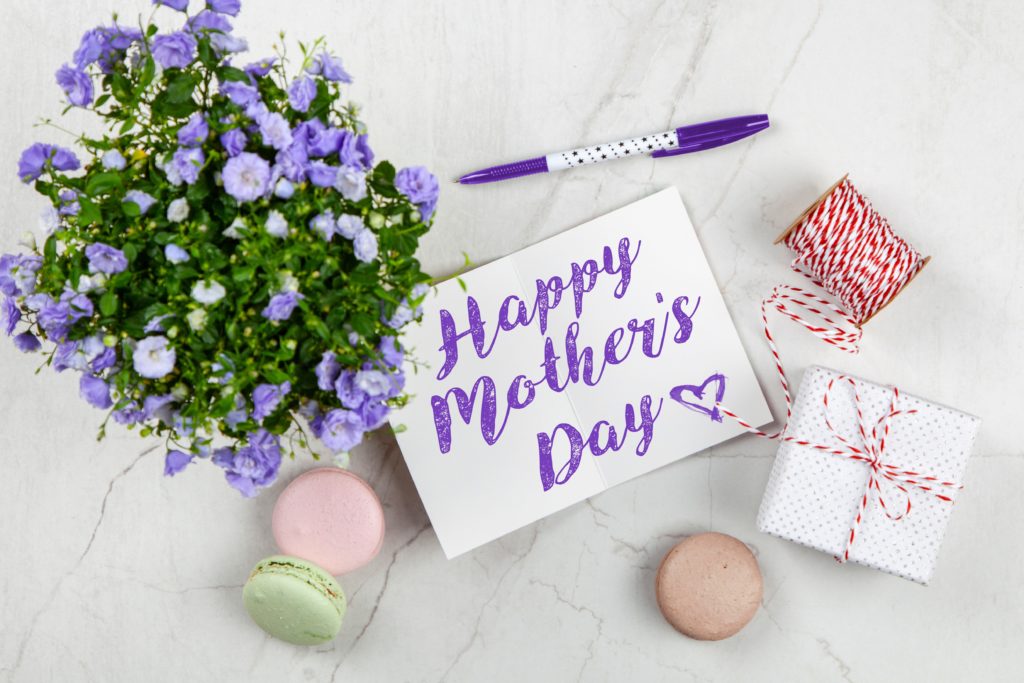 Mother's day gifts: Flat lay on a pale gray marble surface featuring purple flower plant, uncapped purple-and-white pen with a dotted design; white card that says "Happy Mother's Day" in purple script writing with a heart; a spool of red-and-white string; a small gift box wrapped in white polka-dotted paper and tied with the same kind of red-and-white-string as is on the spool; and three macarons in pale pink, pale green, and light brown colors scattered about  