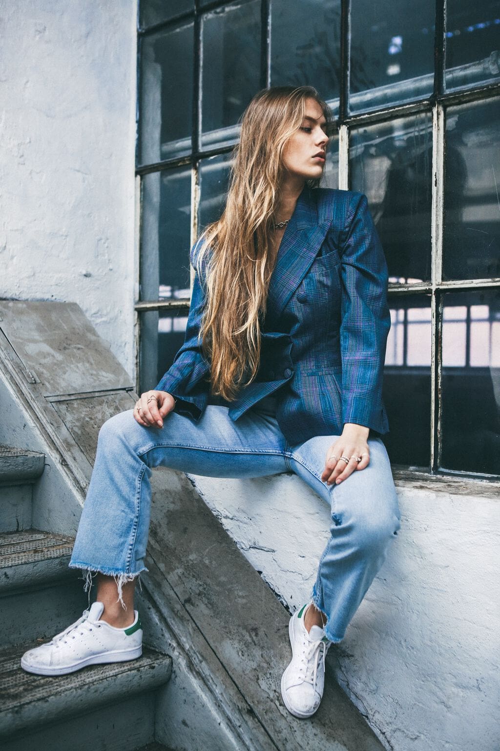 Woman with very long blond hair sitting on a window ledge outside next to stairs, looking away from the camera and wearing a blue plaid double-breasted blazer, loose-fitting light-wash jeans with distressed hems, and Adidas sneakers